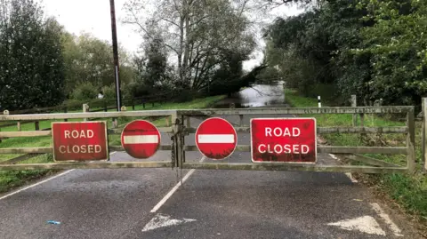 John Devine/BBC The A1123 Earith causeway road, with red 'road closed' and 'no entry' signs in place on a four-bar double gate, padlocked in the middle. The flooded road beyond has a fallen tree across the carriageway and it winds into the distance.