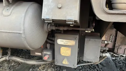 A close-up of a grey metal box underneath a train with SAND CONTROL VALVE written on a gold plaque.