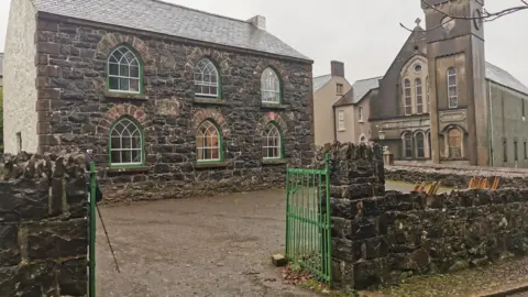 A set of open green gates creates an entrance to a yard, otherwise surrounded by a old stone wall. At the centre of the yard is an old school building, also made of stone. In the background an unadorned chapel, made up of one lower steepled building connected to a tower, looks austere.