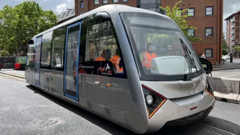 The Very Light Rail tram vehicle on tracks in Coventry city centre. It's a small silver vehicle, similar to the size of a one-deck bus.