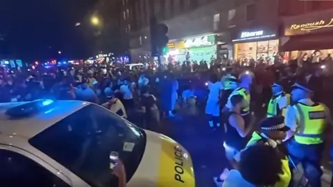 Police stand in the street alongside crowds of people. A police vehicle is in the foreground.