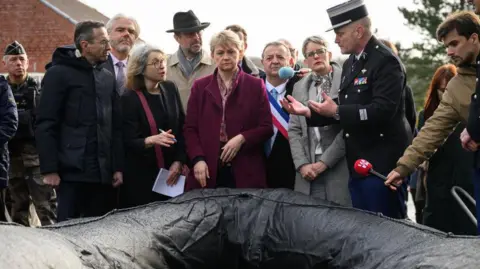 Getty Images Yvette Cooper wearing a purple coat, centre, looks towards the camera at a black dingy. On the left, Bruno Retailleau looks past her to the right at a member of the army in a black coat who speaks to them both. Behind and around them are various other officials and members of the media