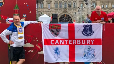 The image shows two men posing with an England flag that has Canterbury written on it. Behind them a castle-esque building can be seen.