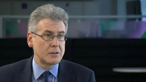 A man with grey hair and glasses in a blue suit and blue shirt against a dark background