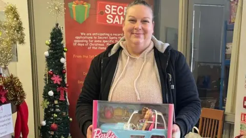 Revive Centre Charity Ann Marie Allsopp holding a Barbie toy. She has a light coloured jumper on and dark jacket. Behind her is a Christmas tree