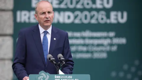 PA Media Taosieach Micheál Martin. He is wearing a navy suit and standing at a green lectern. Behind him is a large green banner with 'IRELAND2026.EU' written on it. 
