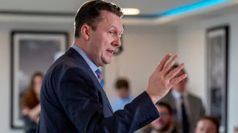 PA Media Russell Findlay, who has short dark hair, speaks with his right hand raised in room full of people 