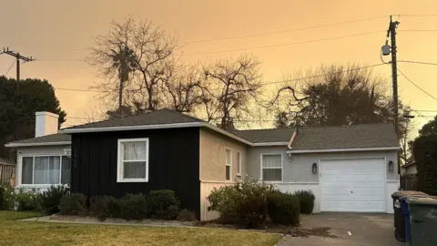 House in Los Angeles with an orange sky behind it, in a road where other properties have been burnt down. The house is one-storey with a lawn and shrubs in the front.