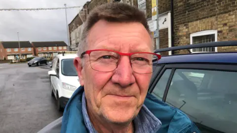 John Devine/BBC Phil Slinger smiles at the camera. He is wearing a blue coat and glasses with a red frame. He is standing in a street with a number of cars parked alongside the pavement behind him. 