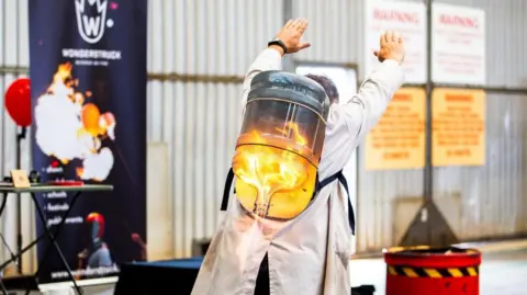 A person wearing a white coat has a flaming rocket contained in an upside down bottle on their back. Their arms are stretched out in front of them. There is a banner behind them which is out of focus but reads WONDERSTRUCK.
