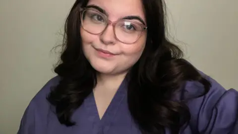Ayla Jones Head and upper body shot of Ayla Jones, who is white and has long dark slightly wavy hair, and is wearing large, light-brown framed glasses. She has a purple medical scrubs top on and is smiling with her head tilted to her left, looking to the left of the camera.