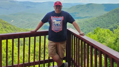 Contributed A man stands on a balcony in front of a large expanse of trees and hills. He wears a navy T-shirt with the Ipswich Town logo on it as well as a red cap.