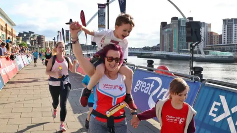 NNP A woman smiles and waves as she reaches the finish line with a young boy on her shoulders