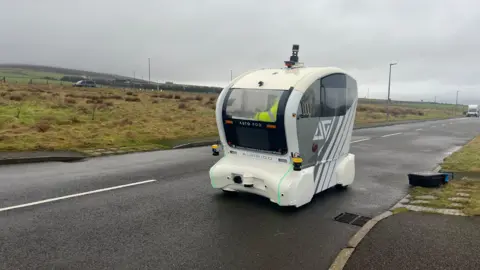 BBC A white vehicle on a road with a sliding door opened and orange lights on the front