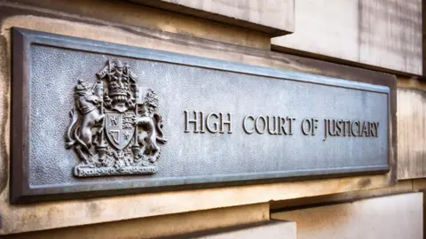 A grey metal sign on an exterior sandstone block wall of the High Court in Edinburgh. The sign has a royal crest and the words "High Court of Justiciary".