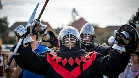 Dave Polier Dos personas vestidas de rojo y negro, con armadura debajo, cascos de metal y espadas, alzando sus armas en señal de victoria.