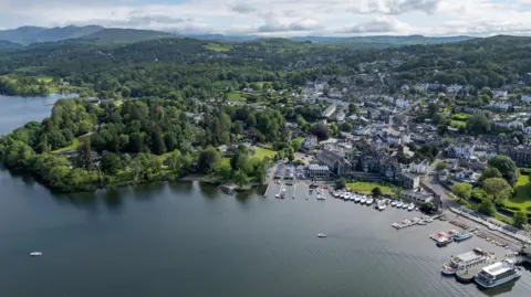 An ariel view of Windermere with the green hills of the Lake District stretching beyond its shore. Lots of boats are tied up and much of the picture is taken up with rows of houses making up a small town amid trees 