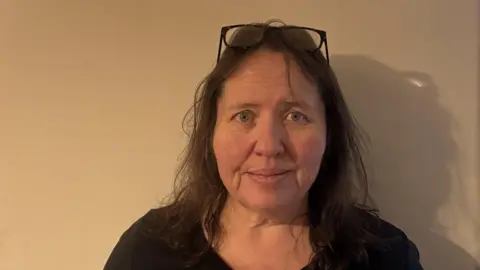 Sarah Cadwallader poses for a photo against a white wall. She has dark hair with glasses perched on top of her head, and wears a black top.