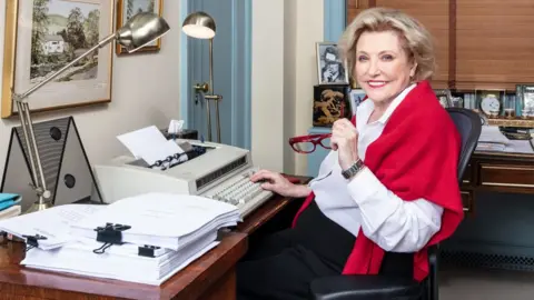 Caroll Taveras/Bradford Enterprises Barbara Taylor Bradford sitting astatine  a typewriter successful  an office. she is wearing a achromatic  garment  and a reddish  shawl and is holding her reddish  glasses successful  her near  hand.