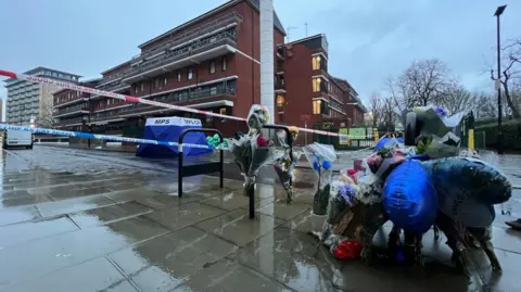 A number of flowers are left at a rainy looking crime scene with a blue MPS tent stationed in the background. Police tape lines the area.