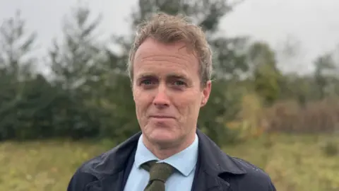 A man stands in a green field wearing a navy coat, blue shirt and green tie. He has short brown hair. 