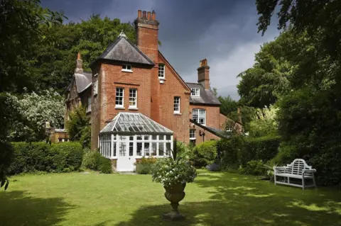 National Trust Images-Andrew Butler The rear view of Max Gate house and garden. The house is a large, detached, Victorian-style house of brick with white-painted sash windows. On the back is a large conservatory. In the foreground is a neat lawn with a planter in the centre and a white Lutyens-style bench on one side.