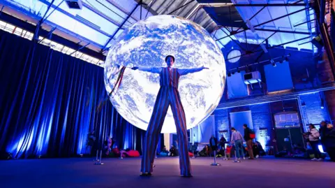 Swindon Borough Council A man on stilts wearing stripey trousers and waistcoat, standing in front of an illuminated moon