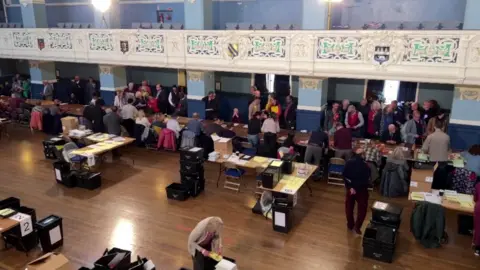An election count at Oxford Town Hall - the image is taken from a balcony looking down at the hall, where there are counters around trestle tables and black ballot boxes.