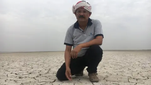 BBC Indian man in turban kneels down on cracked earth covered in a thick layer of white salt