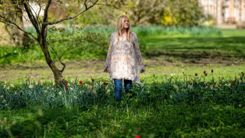 Ben Birchall Sarah Class standing in a patch of flowers wearing a white floral dress and blue jeans