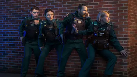Two woman and two men stand with their backs to a brickwall as they turn to their left. The woman at far right partially peers round the corner of the wall, with her right hand up, behind her. All four wear matching green tops and bottoms with a balck police vest.