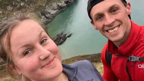 Man and woman smiling at the camera. Man has a red jacket on and rucksack and the woman has a purple top on. The sea is behind them and some steep cliffs.