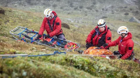 Three search and rescue team members are kneeling on a moor wearing uniform and helmets. One is working with a stretcher while the other two have large backpacks of equipment in front of them. There is a rope attached to one of them which lies in the foreground.