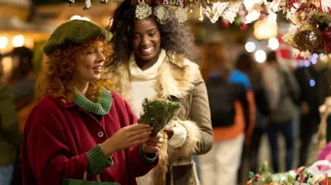 Getty Images Two woman are buying Christmas decorations and gifts at a market, surrounded by festive lights. One of the women is wearing a red coat and a green jumper, and the other woman has a cream jumper and a coat. The market is blurred in the background.