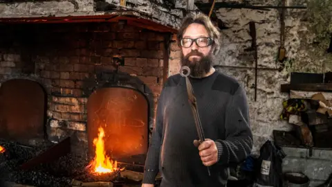 Jill Jennings Rowan Taylor at his forge in Ellesmere. He is holding up a component for a replica of an iron age object. The forge is roaring behind him and he is wearing protective goggles and a thick black jumper.