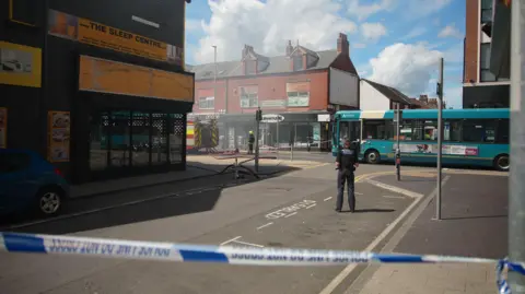 Police tape is barring off a street. Beyond the tape is a female police officer with her back to the camera. The end of a fire engine can be seen behind a shop and the blue sky is marred by smoke.