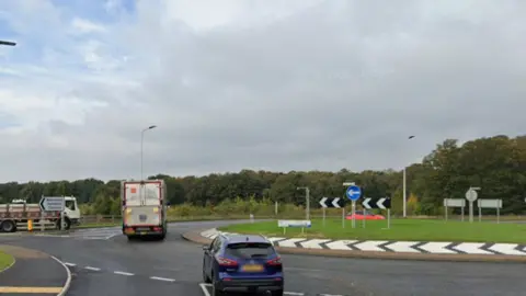 Cars and a lorry appear to be driven around a roundabout which is a grassy area in its centre. It is cloudy.