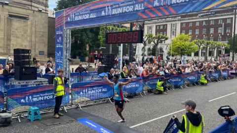 A road race finish line is set up on a city street with a large overhead banner reading “Jane Tomlinson’s Run for All.” A digital race clock suspended above the line displays 00:37:38. A runner in athletic clothing crosses the finish line with one arm raised. Event staff wearing high-visibility vests stand on either side, and a large crowd of spectators gathers behind metal barriers decorated with event branding. In the background, there are trees and multi-storey stone and brick buildings, suggesting an urban setting.