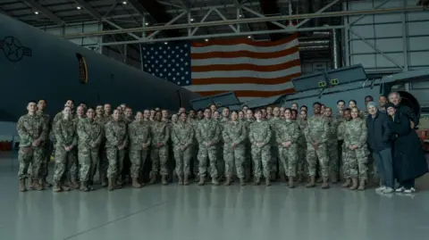 RAF Mildenhall A large group of military personnel stand in a plane wing on an airbase. They all wear matching camouflage uniform and stand with their hands in front of their bodies. 