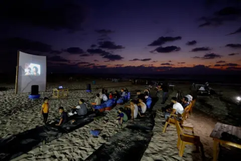 Mohammed Salem / Reuters Families watch a large projector screen at a beachfront café