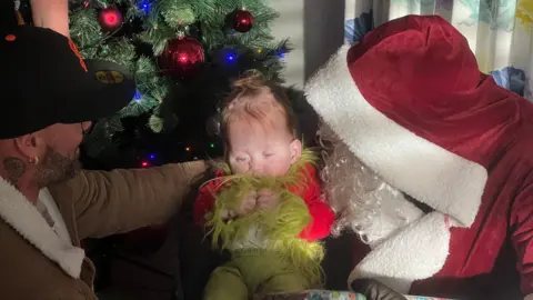 A baby in Grinch outfit sitting on Santa Claus' lap with their dad next to them.