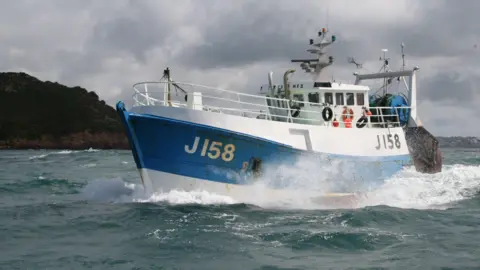 States of Jersey Police A white and blue fishing boat with J158 on the left side, in the ocean, next to another vessel out at sea with grey skies and land seen in the background.