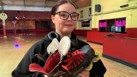 BBC Georgia-Lea has dark brown hair and wears large black-framed glasses. She wears a black hoodie and holds a pair of black and red roller-skates.