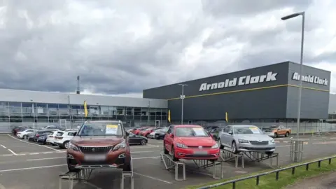 Arnold Clark headquarters in Glasgow. Cars sit in the forecourt and there is a large grey building in the background. 