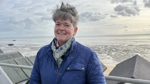 BBC/Tom Larsen-Wright A woman wearing a blue coat standing at Thorpe Bay in Essex smiling at the camera