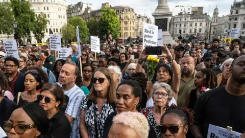 A crowd of people gather in Trafalgar Square as part of a vigil for Jason Arday. Some of them are holding placards.