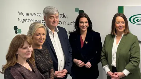 Five people standing side by side. From left to right: Caoimhe Archibald with long brown hair, Michelle O'Neill with blonde hair tied back, Hugh Cormican with short grey hair, Emma Little-Pengelly with long black hair and another lady with shoulder length brown hair.