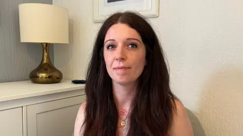 A woman with long brown hair smiles at the camera. She has two red scars on her chest. She sits in a living room with white walls and a white lamp on a table behind her.