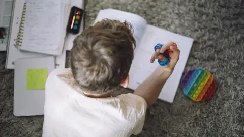 A boy lies on carpet with school books open and with a sensory rubber blue and red toys in his hands. He is facing the floor, away from the camera, and is wearing a white T-shirt.
