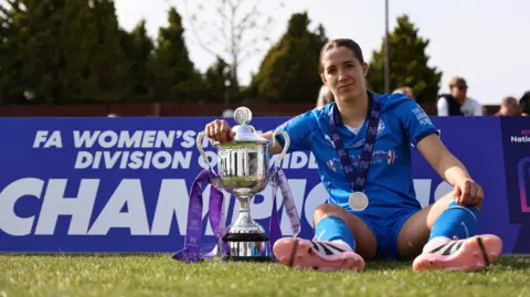 DARREN WILES Poppie Brown sitting on the football pitch with her arm round the trophy. She is wearing a blue Posh kit, football boots and a medal. 